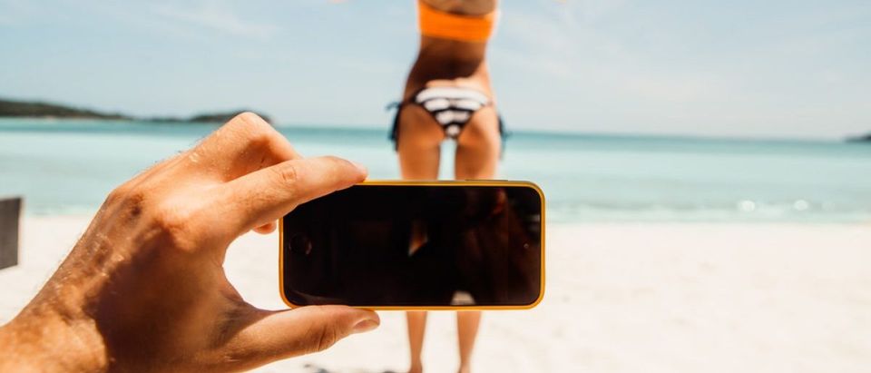 Man taking a photo of a woman in a bikini on the beach. [Shutterstock - Nikkolia]