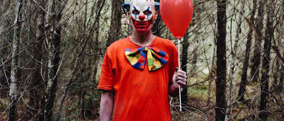 Shutterstock/ a scary evil clown wearing a dirty costume, holding a red balloon in his hand, in the woods (Shutterstock/nito)